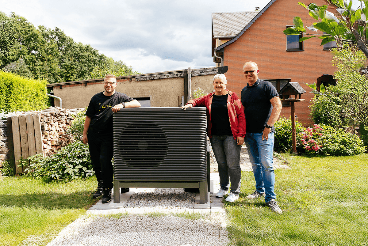 Eine zufriedenes Ehepaar steht mit einem Installateur neben ihrer Wärmepumpe im Garten